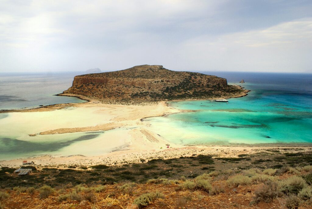 Lagune mit Sandbank und türkisem Wasser auf Kreta, dahinter ein felsiger Hügel im Meer